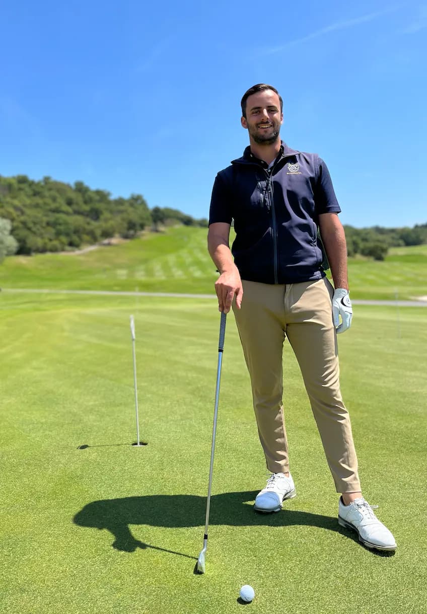 Jules Artale standing on a green, an iron in hand, in front of a fairway and wooded hills.